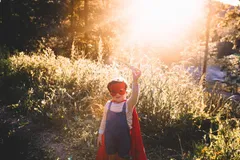 A toddler in a red mask and cape over dungarees stands in long grass in low sun, one arm raised, holding a toy