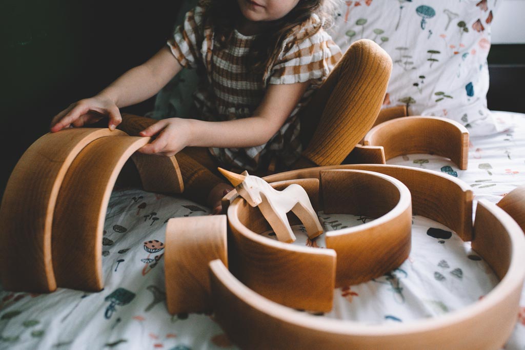 A child arranging Grimm's wooden rainbow blocks into an enclosure on a print-covered bedspread
