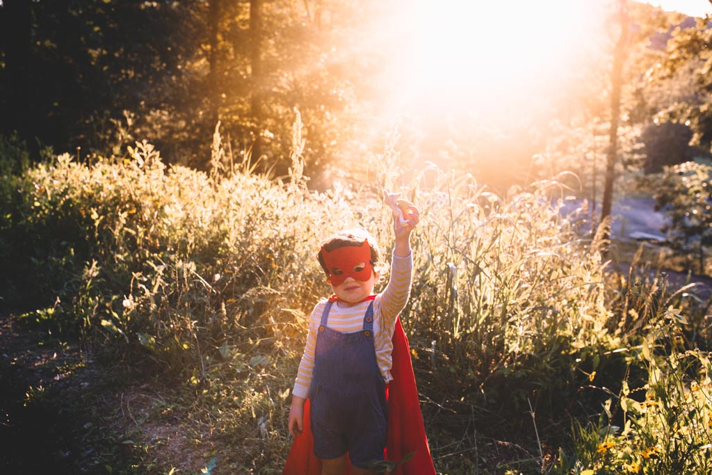 A child in a red mask and cape standing in tall sunlit grass, arm raised