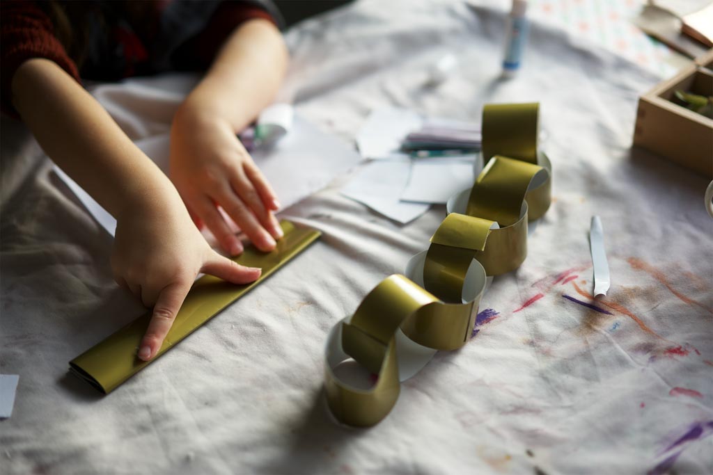 A small hand pressing along a folded gold paper strip beside a half-finished paper chain